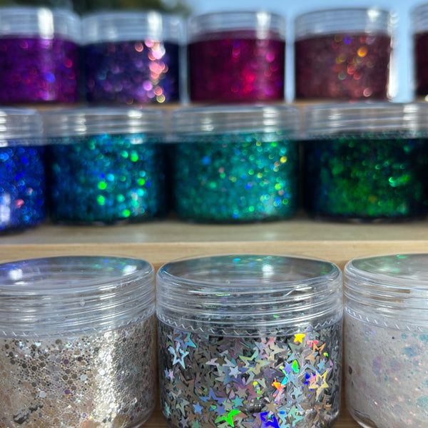 Jars of glitter in various colors on a wooden surface with a blurred outdoor background.