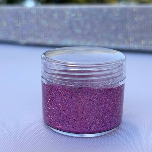 Small jar of dark purple glitter on a textured surface with blurred outdoor background
