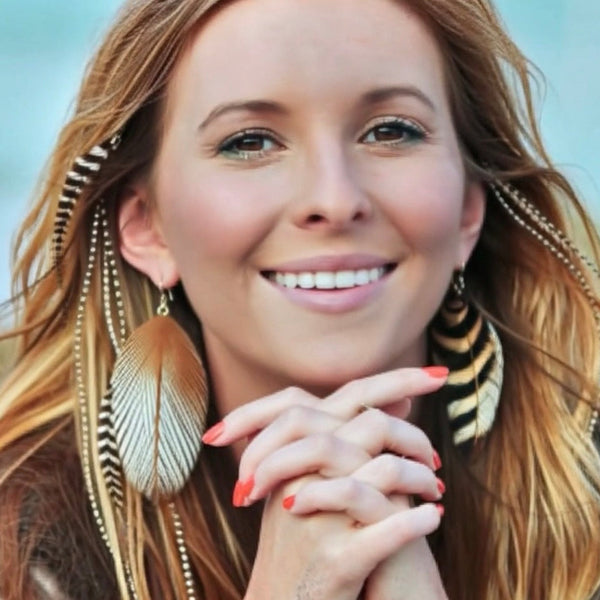 Woman with long blonde hair and feathered earrings smiling outdoors
