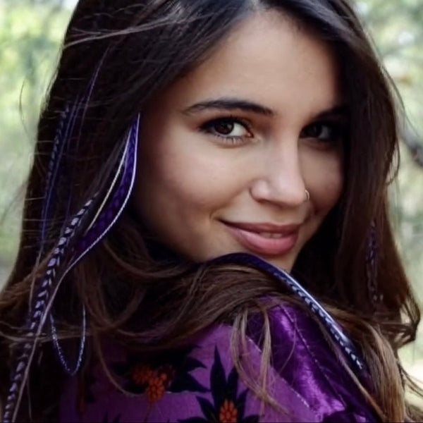 Woman with long brown hair wearing purple feather extensions smiling outdoors.