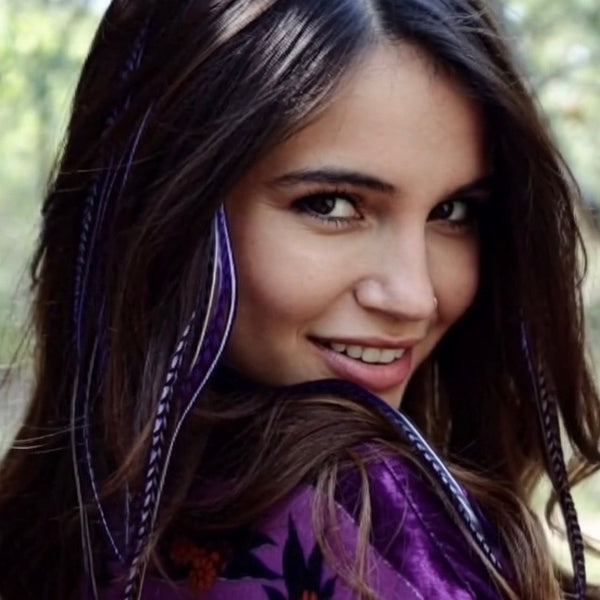 Woman with long brown hair wearing purple feather extensions, with floral pattern top, outdoors.