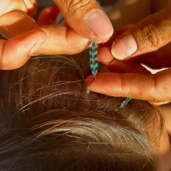 Close-up of hands with blue feather extension attaching into hair with nano bead, with a blurred background.